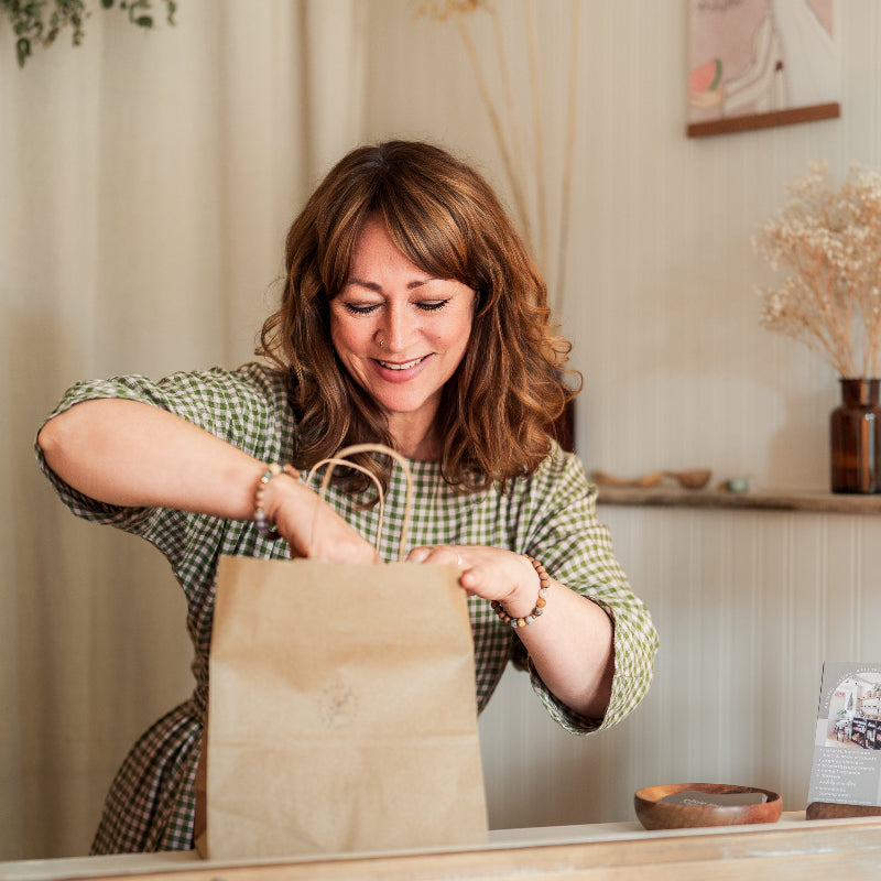 Woman in a checkered shirt opening a brown paper bag in a cozy indoor setting.