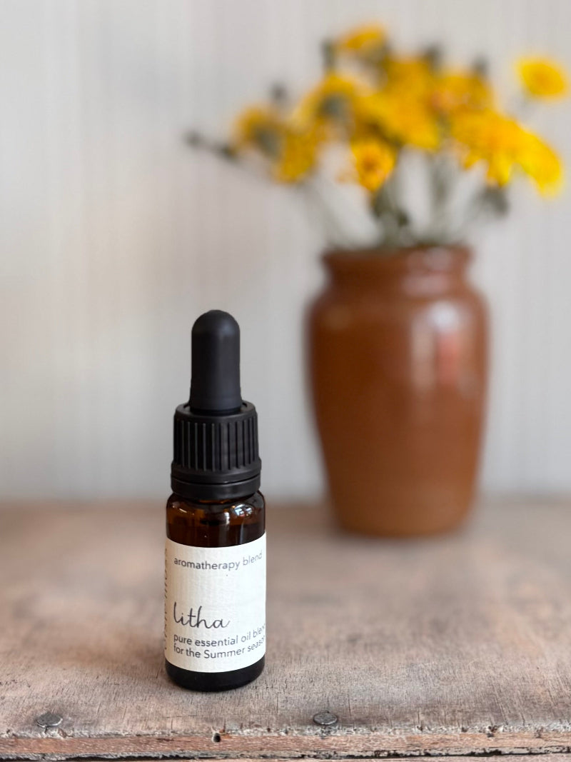 Bottle of Litha essential oil with a dropper on a wooden surface with a vase of yellow flowers in the background.