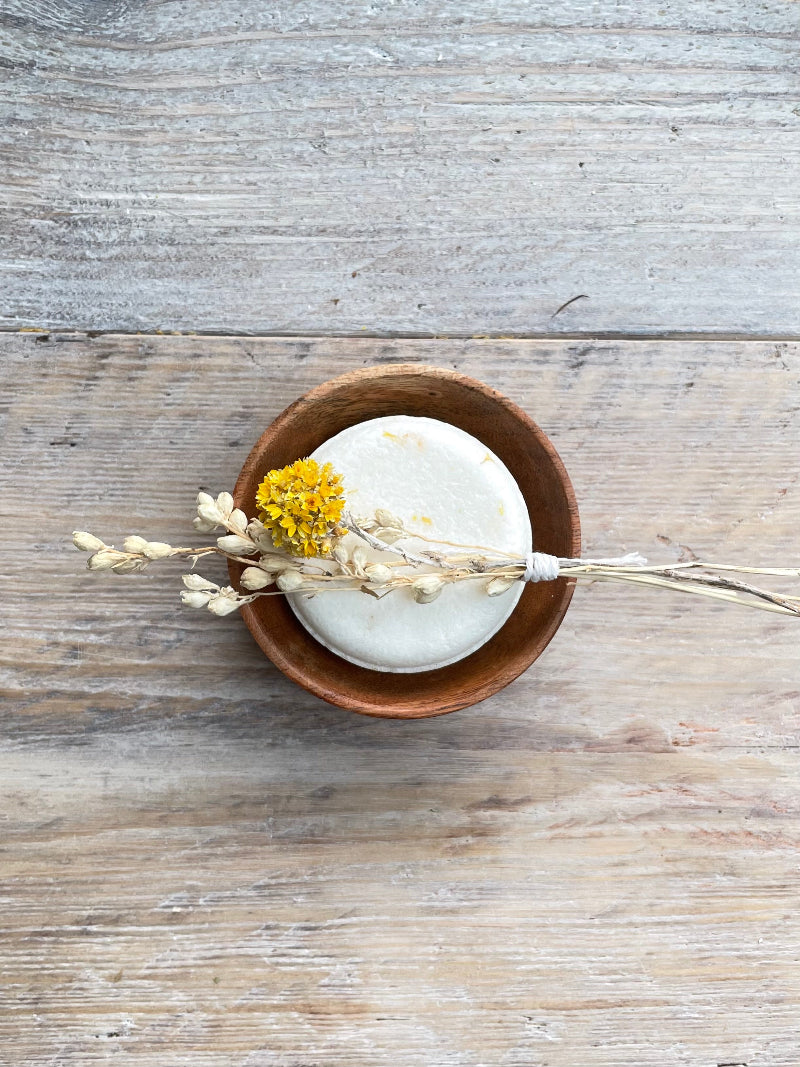 White shampoo bar in a wooden bowl with dried flowers on a wooden surface