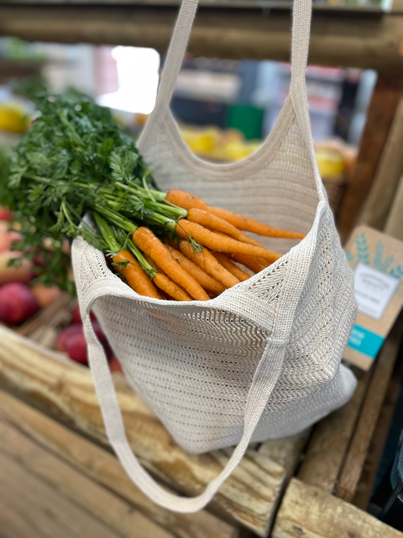 A beige antique lace bag filled with carrots, displayed on a wooden rack with various fruits and vegetables in the background.