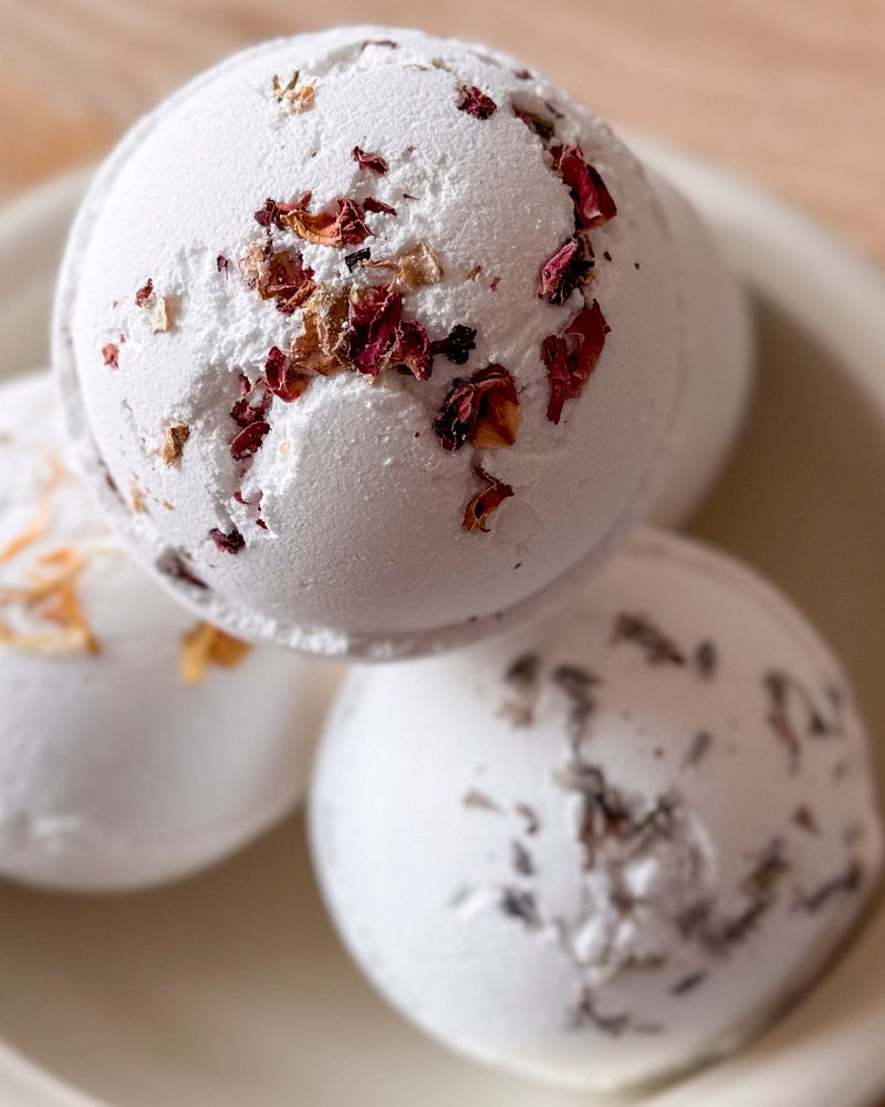 Three white bath bombs topped with dried botanicals, arranged in a shallow beige dish on a light wooden surface.
