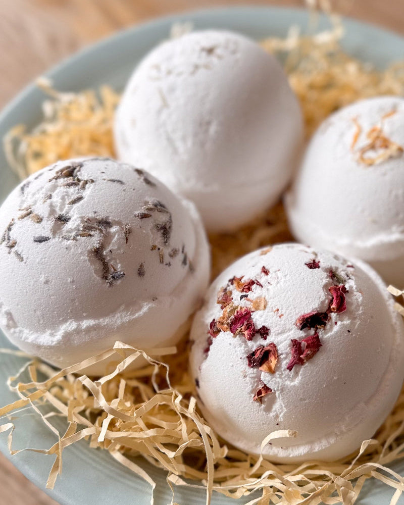 Four white bath bombs with dried botanical inclusions, arranged on a light blue plate with shredded paper filler on a wooden surface.