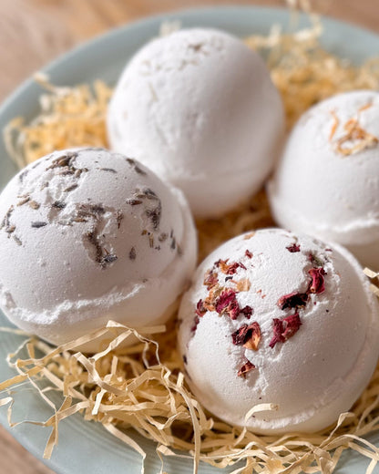 Four white bath bombs with dried botanical inclusions, arranged on a light blue plate with shredded paper filler on a wooden surface.