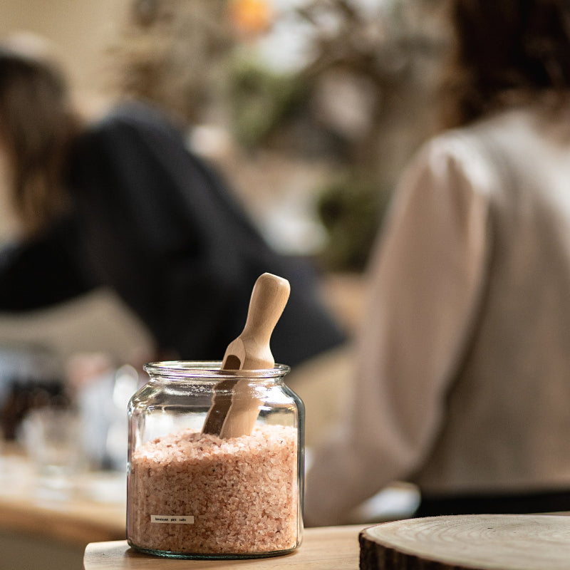 Glass jar with pink salt and wooden spoon on a blurred background of people sitting at a table.