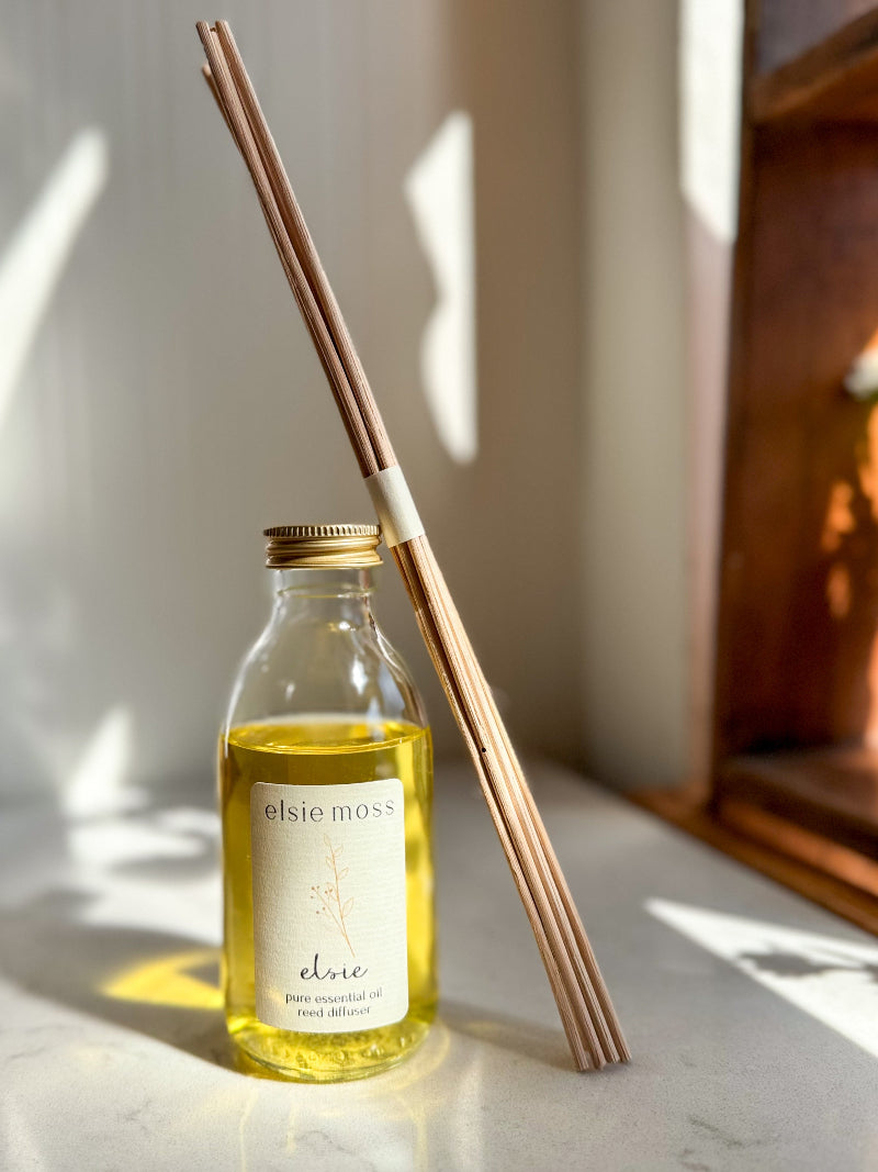 A clear glass bottle of Elsie Moss Chloe pure essential oil with a gold cap, next to four natural bamboo reed diffuser sticks, set on a marble surface with warm sunlight and shadows.