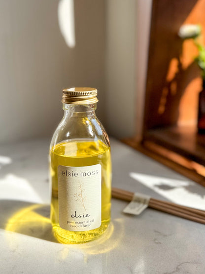 A clear glass bottle of elsie moss elne pure essential oil with a gold cap and cream label, sitting on a marble surface with warm sunlight and shadows.
