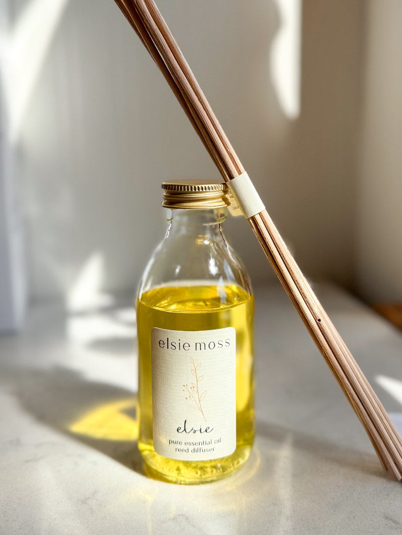 A clear glass bottle of Elsie Moss Elmo pure essential oil with a gold cap and white label, filled with yellow oil, standing on a light marble surface. A wooden reed diffuser stick rests diagonally across the bottle against a softly lit neutral background.