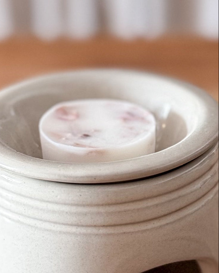 A small ceramic wax warmer with a matching lid and a round wax melt inside, set on a wooden tabletop with a softly blurred background.