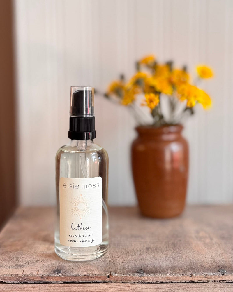 Elsie Moss letha room spray bottle on a wooden surface with a vase of yellow flowers in the background.
