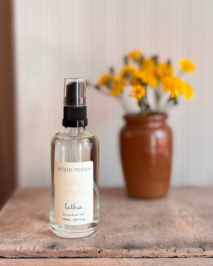 Elsie Moss letha room spray bottle on a wooden surface with a vase of yellow flowers in the background.