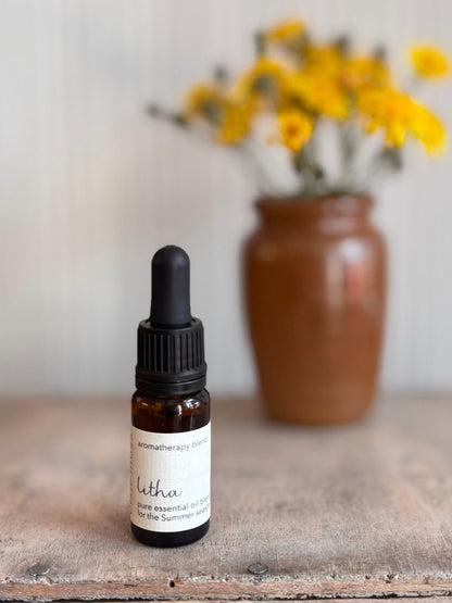 Bottle of Litha essential oil with a dropper on a wooden surface with a vase of yellow flowers in the background.
