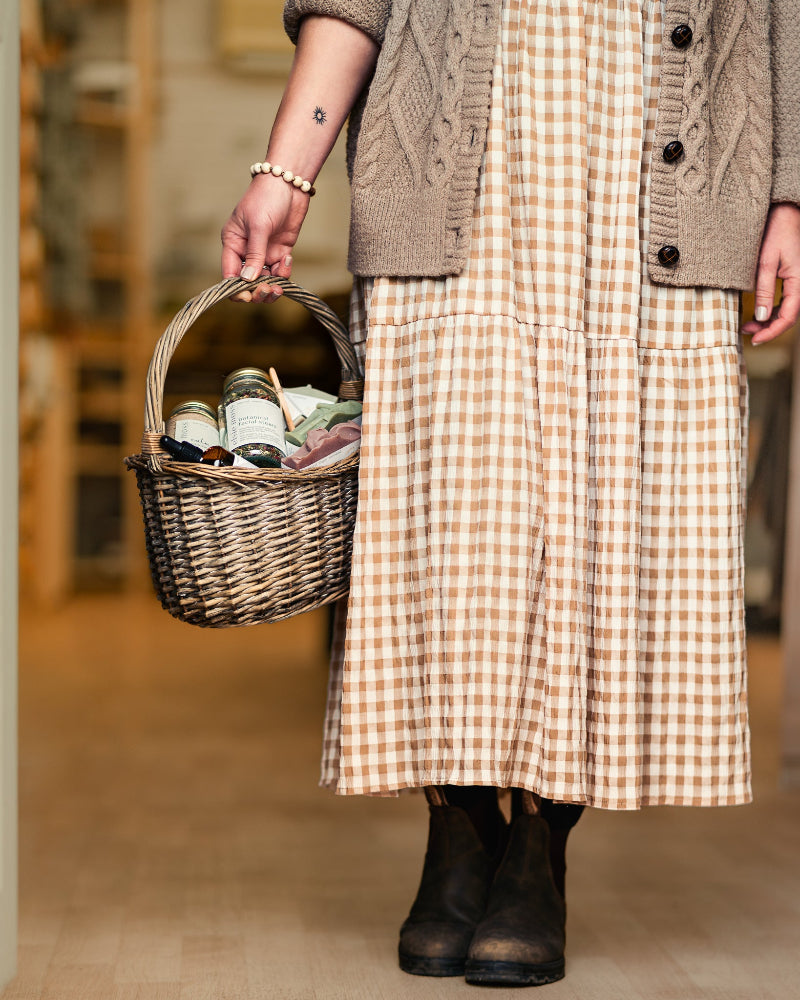 Person wearing a checkered dress and cardigan holding a woven basket with items.