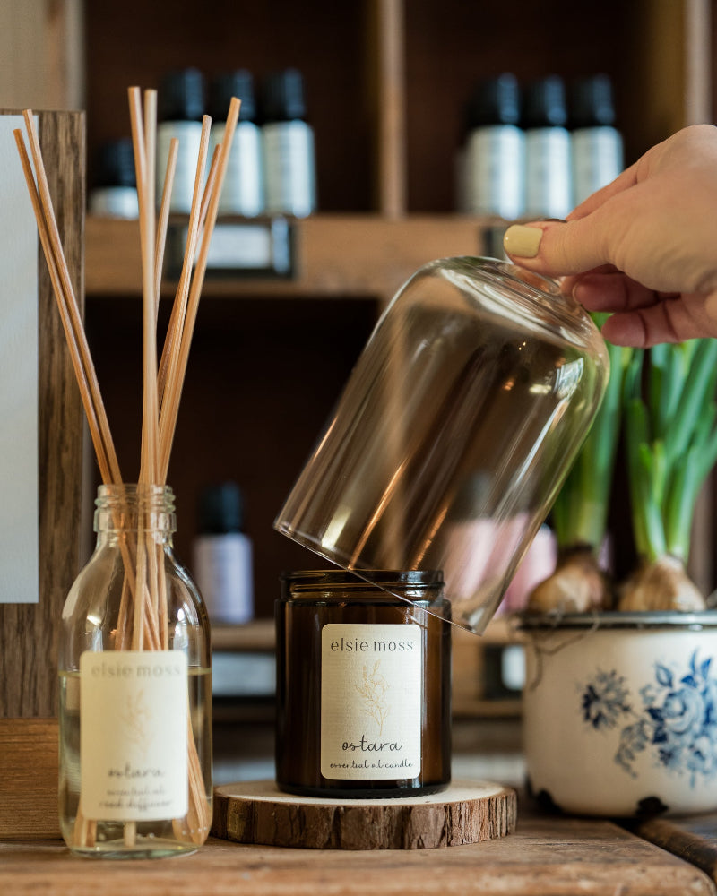Hand pouring a liquid into a glass bottle with reed diffuser and candle on a wooden surface.