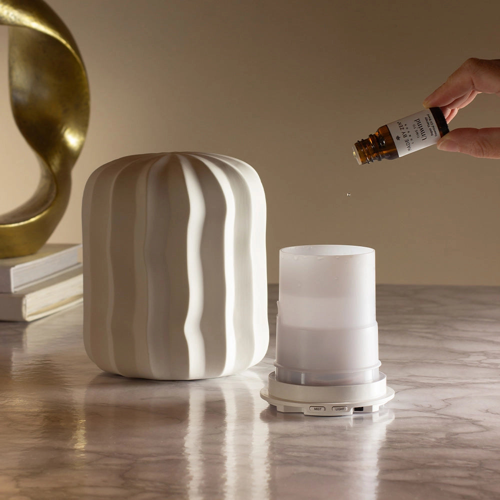A white ribbed diffuser container sits on a marble surface next to its translucent top chamber, with a hand holding a small amber bottle above it. A gold abstract sculpture and stacked books are visible in the background against a warm beige wall.