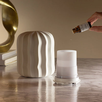 A white ribbed diffuser container sits on a marble surface next to its translucent top chamber, with a hand holding a small amber bottle above it. A gold abstract sculpture and stacked books are visible in the background against a warm beige wall.