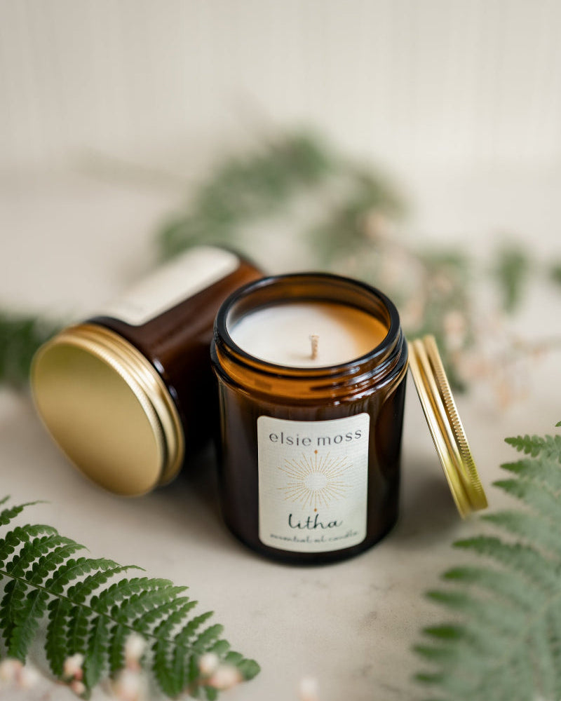 Candle in a glass jar with a visible brand label, surrounded by greenery on a neutral background