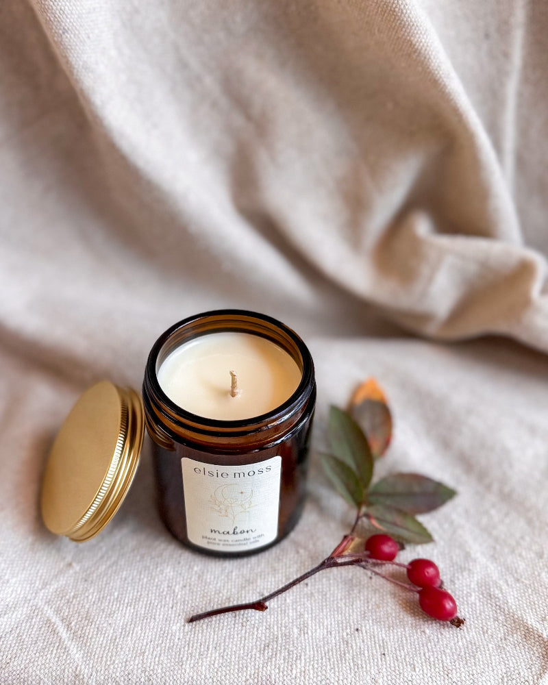 An open amber glass candle jar with cream wax and a gold lid resting beside it on a beige fabric surface, styled with a small branch and red berries. The label reads &