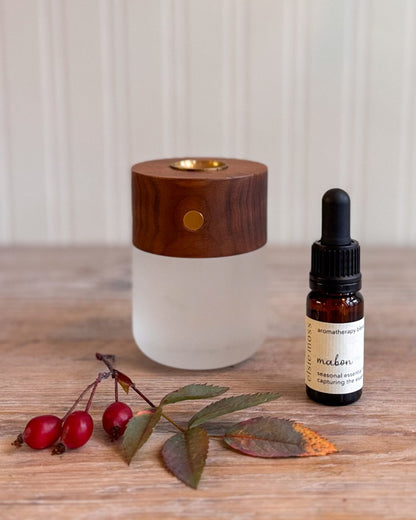 A diffuser and essential oil bottle styled on a wooden surface with red berries and leaves. The diffuser has a frosted white glass body and a dark wood top with a brass accent.