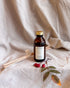 An amber glass bottle of Elsie Moss Mabon essential oil reed diffuser with a gold cap and white label, styled on a beige linen fabric with wooden reed diffusers and rosehips.