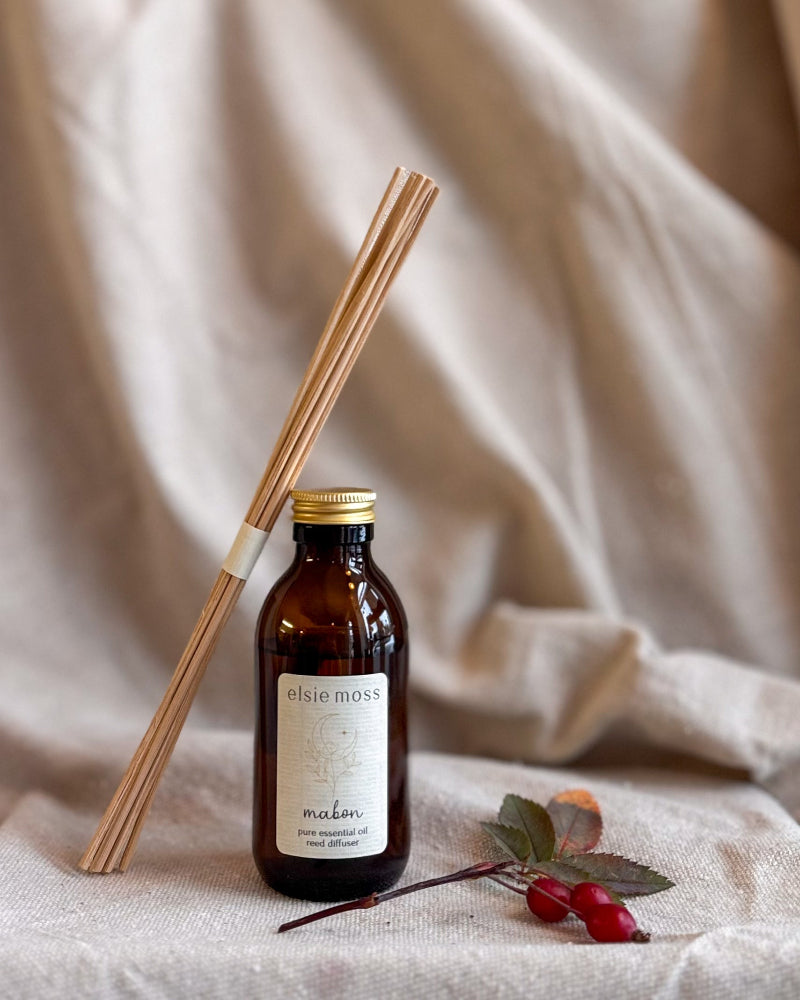 An amber glass bottle of elsie moss essential oil diffuser with a gold cap and a bundle of natural wood reeds, styled on a beige linen fabric with rosehips.