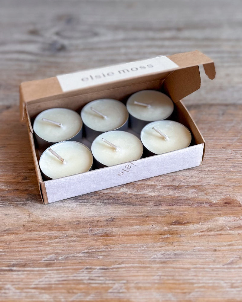 Six small cream-colored tea light candles in a brown cardboard box with a white paper label reading &