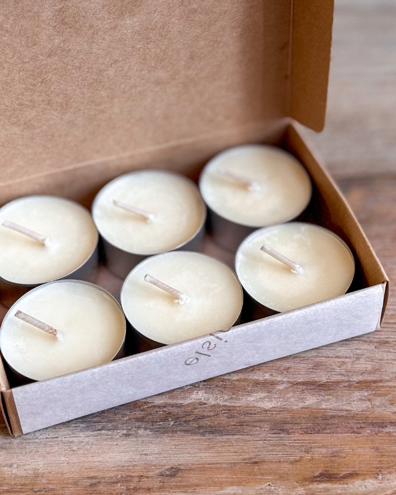 Six small cream-colored tea light candles in a brown kraft gift box, set on a rustic wooden surface.
