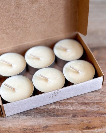 Six small cream-colored tea light candles in a brown kraft gift box, set on a rustic wooden surface.