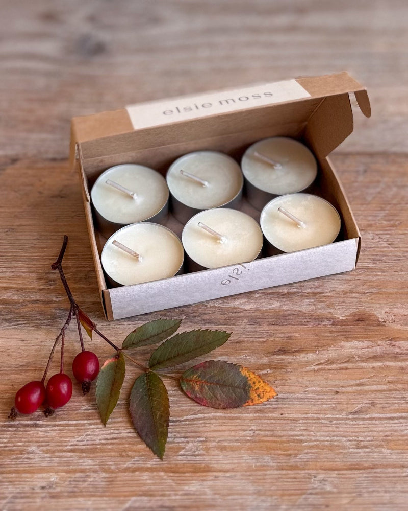 A box of six tea light candles on a rustic wooden table, styled with red berries and green leaves. The brand name &