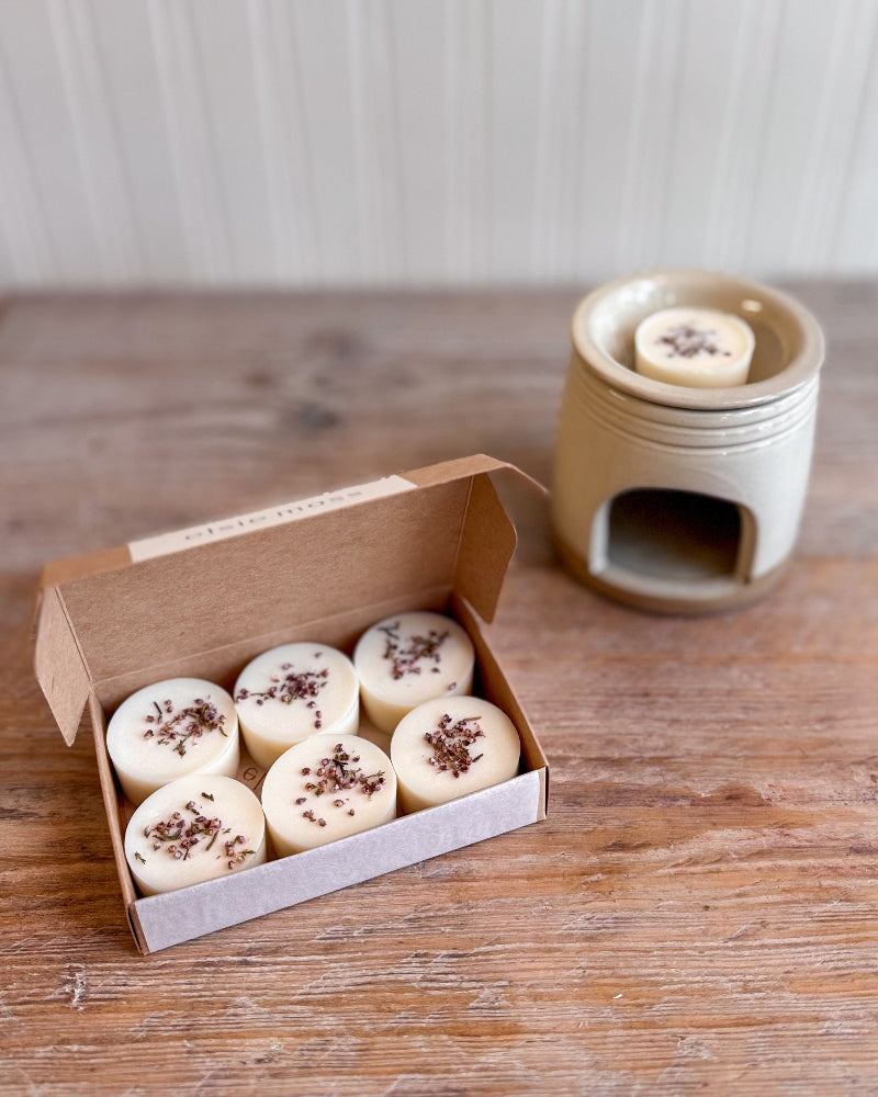 Six round cream-colored wax melts with dried botanicals on top, displayed in an open kraft paper box next to a ceramic wax warmer on a rustic wooden table.
