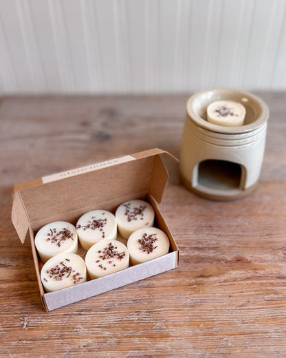 Six round cream-colored wax melts with dried botanicals on top, displayed in an open kraft paper box next to a ceramic wax warmer on a rustic wooden table.