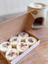 A box of round white wax melts topped with dried flower petals, displayed on a wooden surface with a ceramic candle holder in the background.