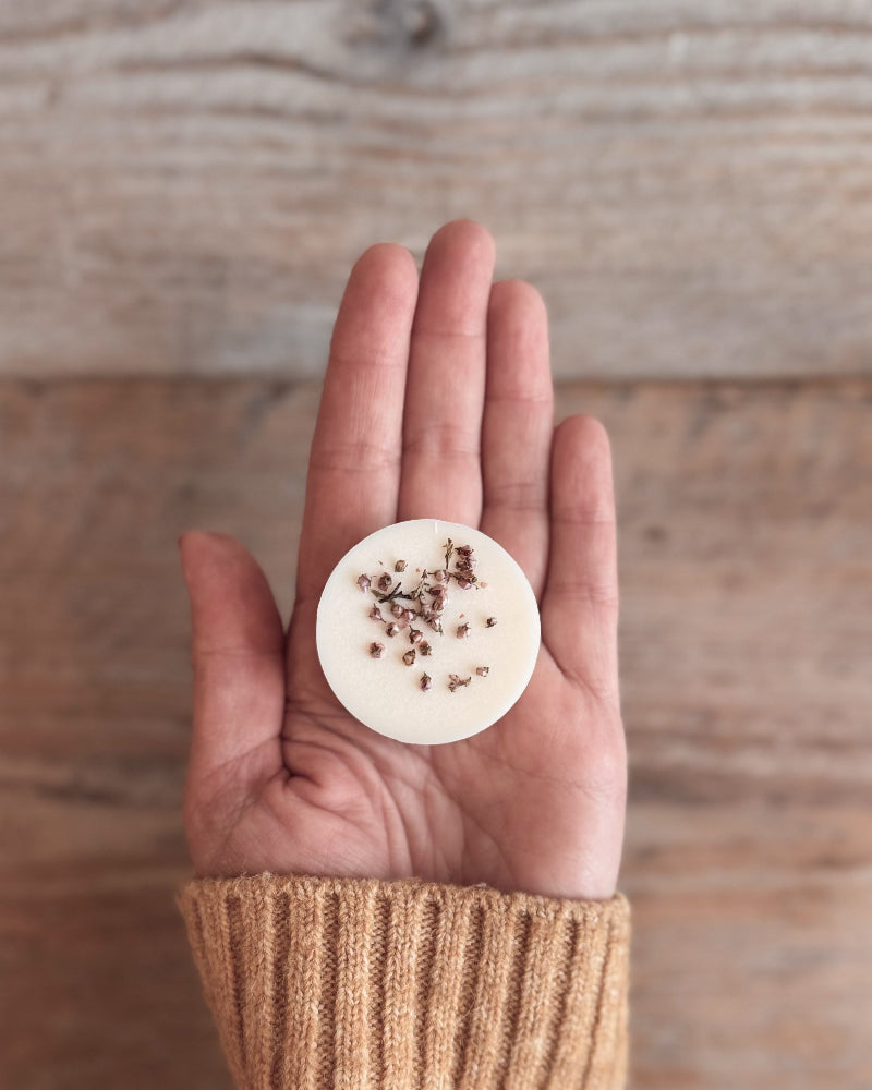 A hand wearing a beige knit sweater holds a small round white wax melt or soap piece topped with dried botanicals against a rustic wooden background.