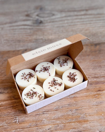 Six round cream-colored wax melts or tea lights in an open kraft paper box, each topped with dried flower petals, displayed on a rustic wooden surface. The box lid is printed with the brand name &