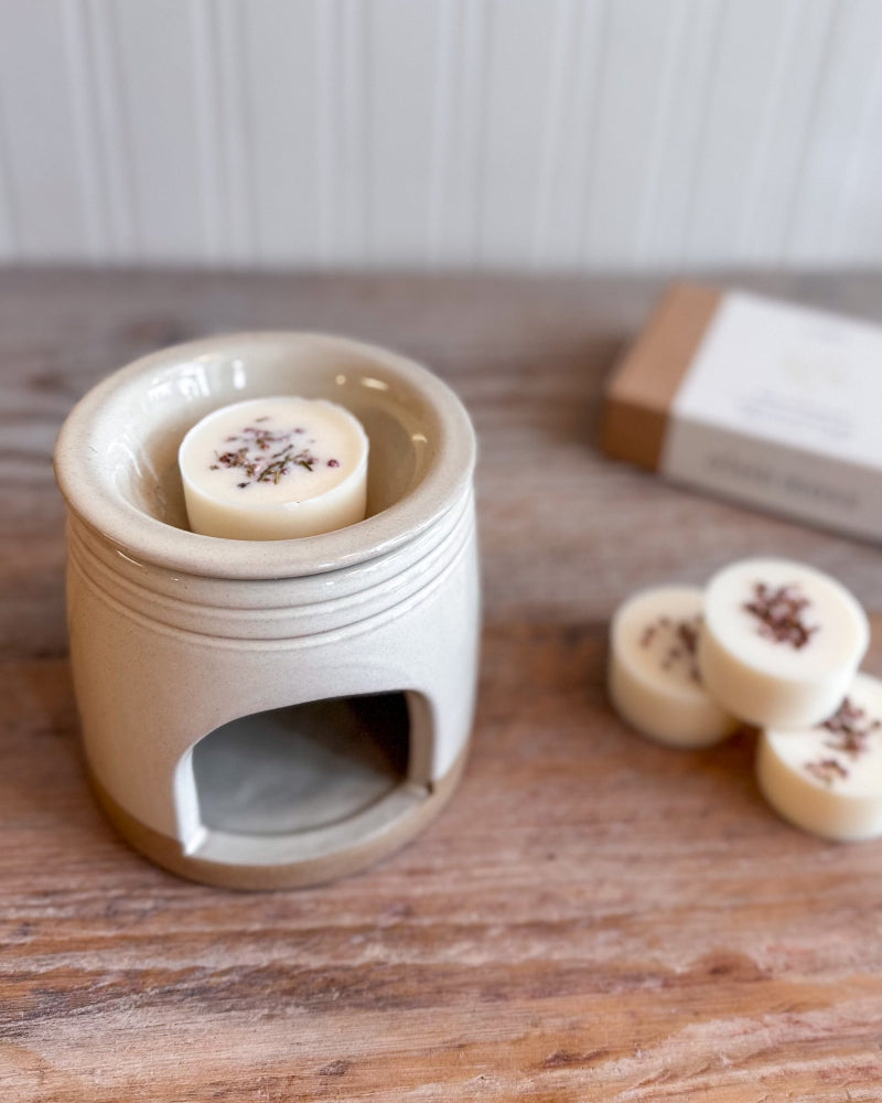 A small ceramic wax warmer holding a round cream-colored wax melt with dark botanical flecks, set on a wooden surface with additional wax melts and a box in the background.
