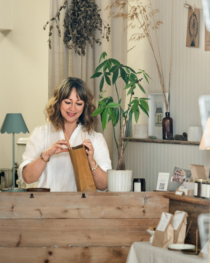 Woman standing behind a wooden counter in a store setting with plants and decor.