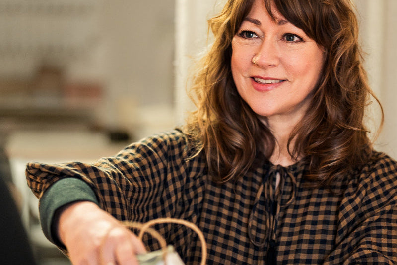 Woman in a checkered shirt opening a brown paper bag with a blurred indoor background