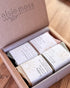 An open kraft gift box from elsie moss BOTANICAL containing four wrapped handmade soaps, photographed on a wooden surface.