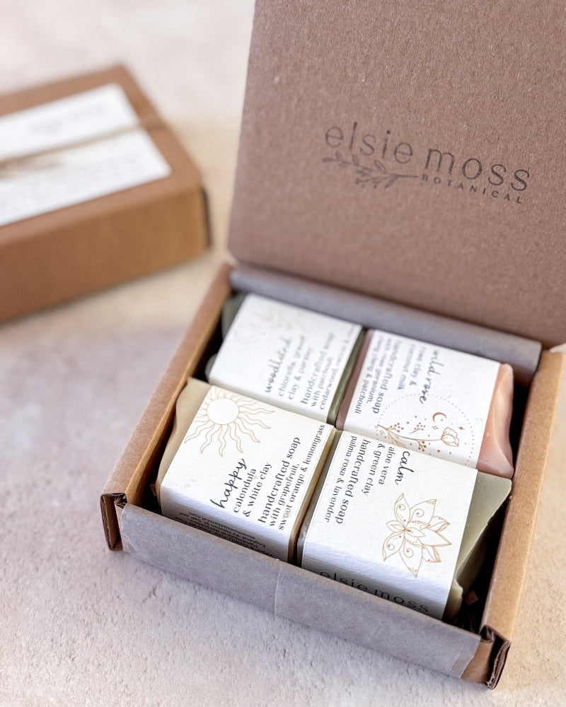An open kraft gift box from elsie moss Botanical containing four wrapped handmade soaps, with one soap bar visible outside its wrapper. The box lid is printed with the brand name &