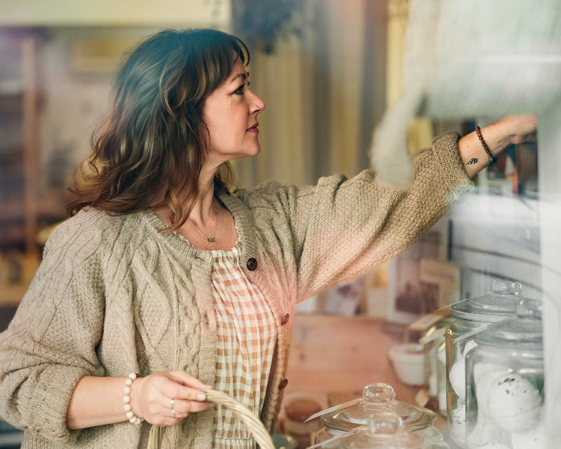 Woman in a store holding a basket with products, surrounded by jars and shelves.