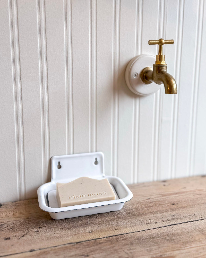 White soap dish with a bar of soap on a wooden surface, next to a gold faucet on a white paneled wall.