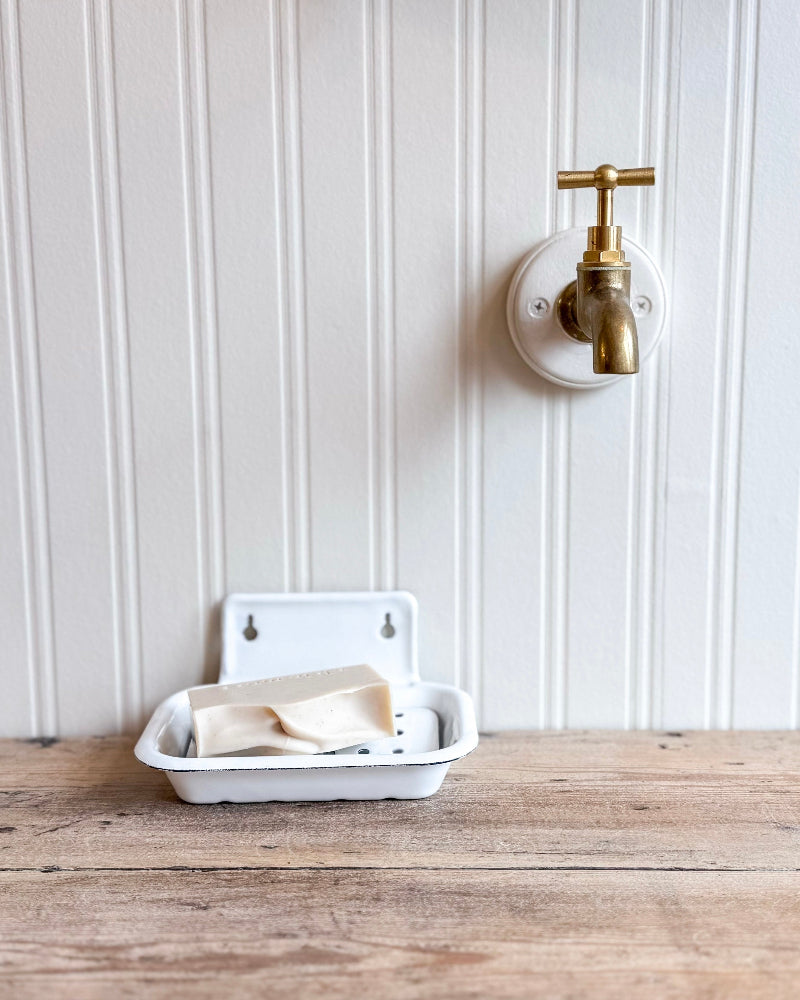 White soap dish with a bar of soap on a wooden surface next to a brass wall-mounted faucet.